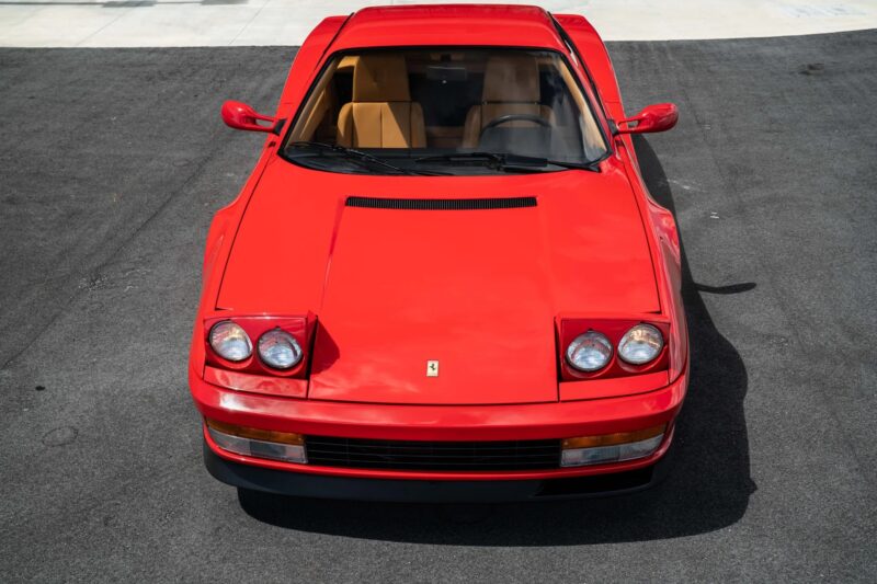 A rare car, this red Ferrari Testarossa with pop-up headlights is parked on a gray asphalt surface, viewed from above.