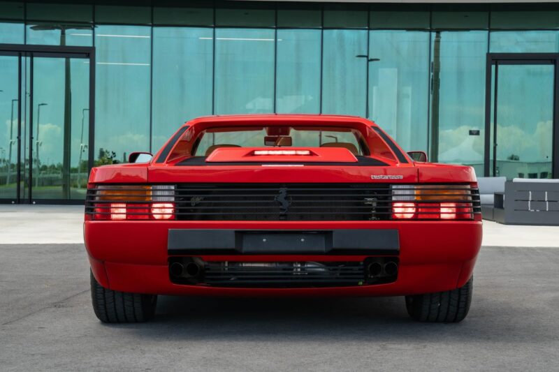 Rear view of a rare sports car—a red 1990 Ferrari Testarossa—parked on pavement in front of a modern glass building.