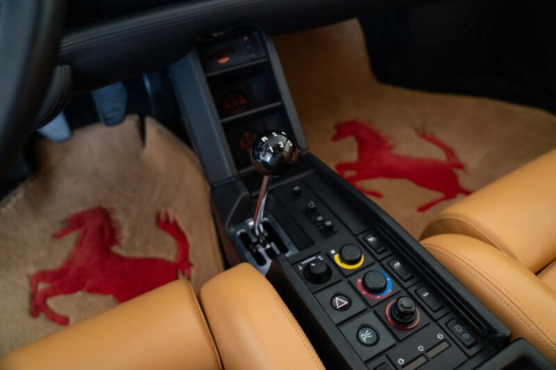 Interior view of a rare car: the 1990 Ferrari Testarossa, featuring a manual gear shifter, control buttons, tan leather seats, and floor mats with red prancing horse logos.