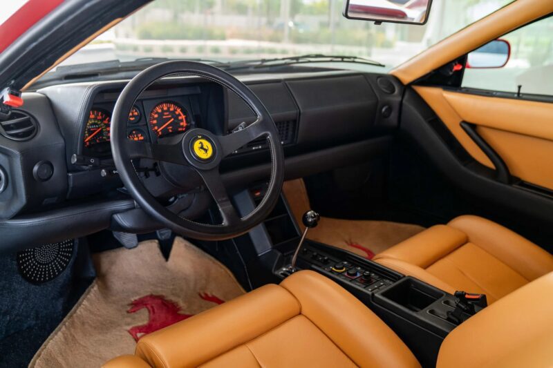 Interior view of a rare Ferrari Testarossa sports car showing a black steering wheel with Ferrari logo, tan leather seats, manual gear shift, and analog dashboard gauges.