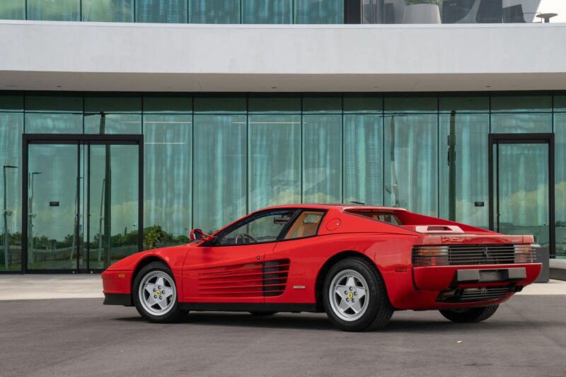 A rare Ferrari Testarossa sports car in striking red is parked on a paved surface in front of a modern glass building with large windows and doors.