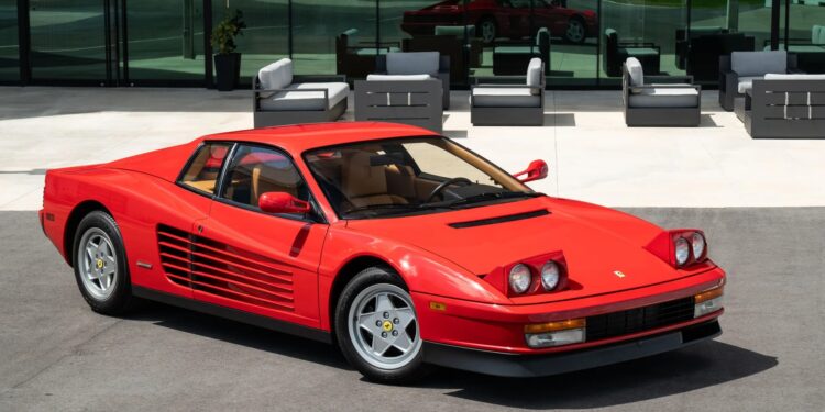 A rare 1990 Ferrari Testarossa sports car is parked on pavement in front of a modern glass building with outdoor seating.