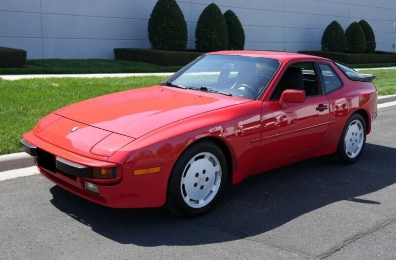 A red Transaxle Porsche 944 coupe is parked on the street near a sidewalk and landscaped greenery, with a modern building in the background—an unsung classic that car enthusiasts may want to invest in.