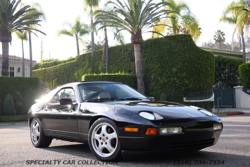 A black Porsche 928 sports car, one of the unsung classics from the Transaxle Porsche era, is parked on a sunny residential street, surrounded by palm trees and hedges—a smart invest for any enthusiast.