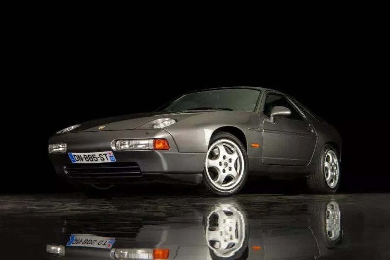 A grey Porsche 928 Transaxle sports car, one of the unsung classics, is parked on a reflective surface against a black background, viewed from the front left angle—a smart choice for those looking to invest in automotive history.