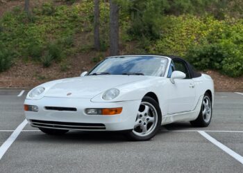 A white Porsche 968 convertible, one of the unsung classics among Transaxle Porsche models, is parked at an angle in an empty outdoor lot with trees and greenery in the background—an ideal car to invest in.