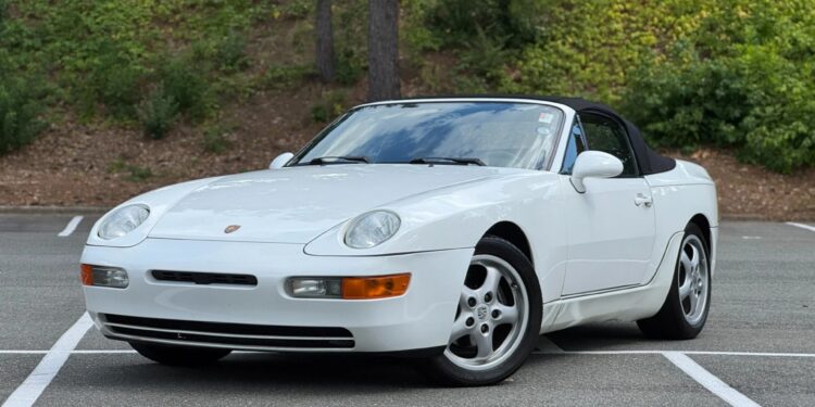 A white Porsche 968 convertible, one of the unsung classics among Transaxle Porsche models, is parked at an angle in an empty outdoor lot with trees and greenery in the background—an ideal car to invest in.