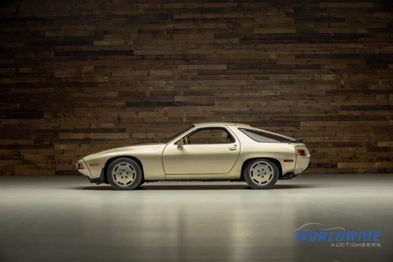 Side view of a cream-colored classic sports car parked indoors on a white floor, set against a wooden plank wall and "Worldwide Auctioneers" logo—an elegant highlight from their diverse auction lineup.