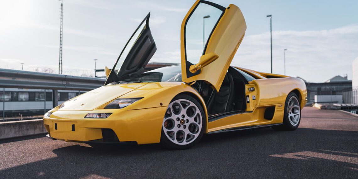 A gleaming yellow Lamborghini Diablo VT 6.0, with its scissor doors dramatically open, rests on a paved surface under the clear blue sky—an icon ready for the spotlight at Broad Arrow Auctions.
