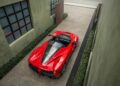 A 2017 Ferrari LaFerrari Aperta sits parked between two green buildings, viewed from above. This red sports car boasts a sleek design and a transparent engine cover that catches the eye, showcasing its elegance and power.