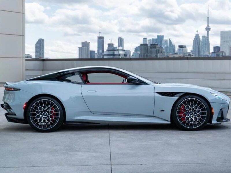A silver Aston Martin DBS 770 Ultimate sports car parked on a rooftop with a city skyline and cloudy sky in the background, exuding grand touring elegance.