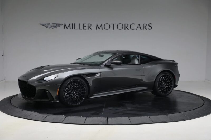 A dark gray Aston Martin DBS Ultimate sports car displayed in a showroom with "Miller Motorcars" branding on the wall behind it, epitomizing the essence of grand touring luxury and performance.