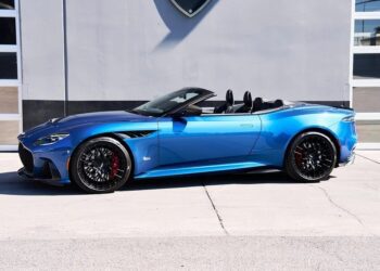 A blue Aston Martin DBS 770 Ultimate Grand Touring masterpiece is parked in front of a gray building with large windows and a garage door.