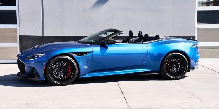 A blue Aston Martin DBS 770 Ultimate Grand Touring masterpiece is parked in front of a gray building with large windows and a garage door.