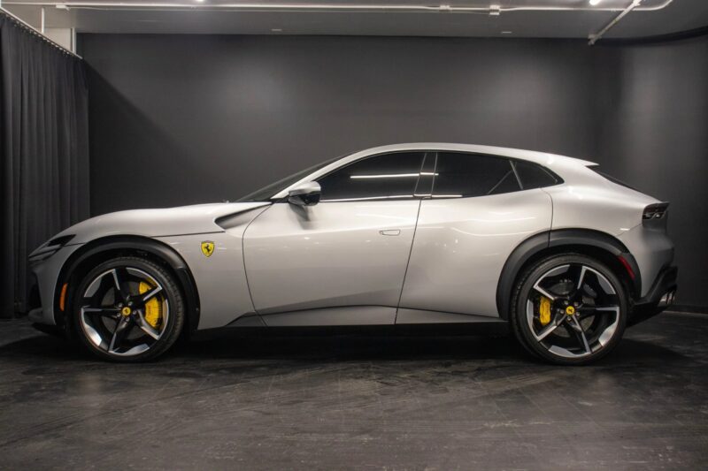 A silver Ferrari Purosangue SUV with black wheels and yellow brake calipers is parked indoors against a plain black wall—an exceptional SUV for sale for those looking to buy Ferrari's latest masterpiece.