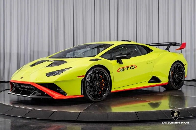 A bright yellow Lamborghini Huracán STO luxury car is displayed indoors on a reflective circular platform, with a gray curtain backdrop.