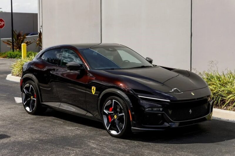 A black Ferrari Purosangue SUV for sale is parked on a paved surface near a beige wall and some greenery.