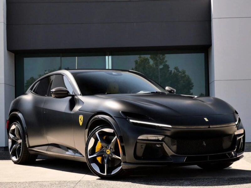 A matte black Ferrari Purosangue SUV is parked in front of a modern building, featuring yellow brake calipers and Ferrari badges on the fender.