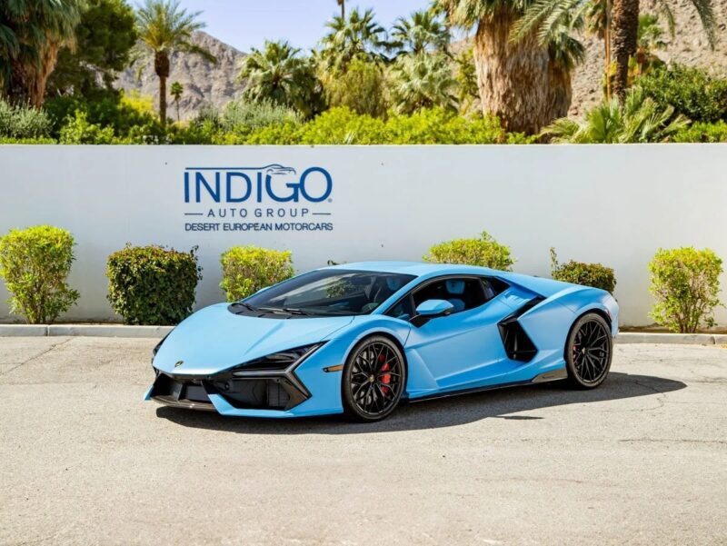 A light blue Lamborghini Revuelto supercar is parked in front of a white wall with an INDIGO Auto Group sign and palm trees in the background.