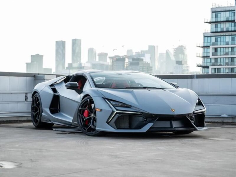 A silver Lamborghini Revuelto supercar is parked on a rooftop with a city skyline and modern buildings visible in the background.