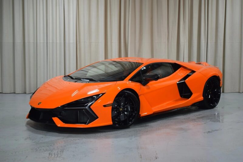 An orange Lamborghini Revuelto sports car is parked indoors on a gray floor, with beige curtains in the background—a stunning example of supercars for sale.