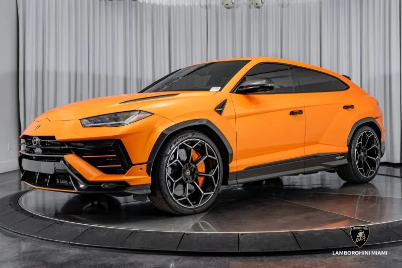 An orange Lamborghini Urus luxury car is displayed in a showroom with white curtains and a Lamborghini Miami sign on the floor.