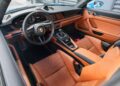 Interior of a Porsche 911 featuring a black steering wheel, touchscreen display, brown leather seats, and manual gear shift. For sale now, this luxury sports car combines sleek design with unparalleled performance—an exclusive driving experience in every detail.