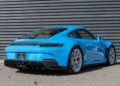 A Riviera Blue Porsche 911 sports car with a spoiler, rear diffuser, and crisp white rims is parked near a gray corrugated metal wall. This sleek beauty is now for sale.