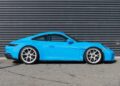A stunning Riviera Blue Porsche 911 with striking white wheels is parked on a paved surface in front of a corrugated metal wall, awaiting its next adventure. For sale now.