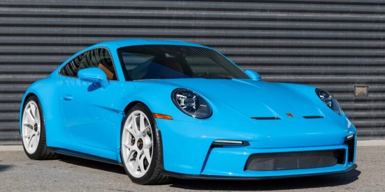 A stunning Porsche 911 S/T in striking PTS Riviera Blue, complete with white wheels, is parked gracefully on a concrete surface, set against a dark, ridged metallic wall. This eye-catching sports car is available for sale to the discerning enthusiast.