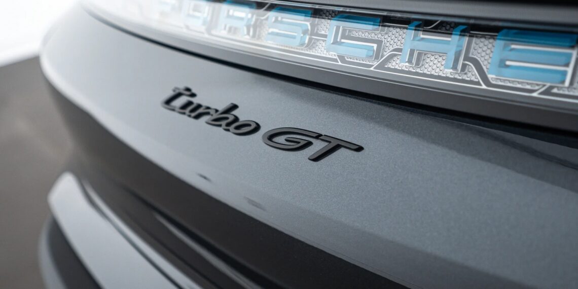 Close-up of the rear badge on a luxury car, highlighting the "turbo GT" badge and part of the illuminated "Porsche" emblem on a metallic gray surface.