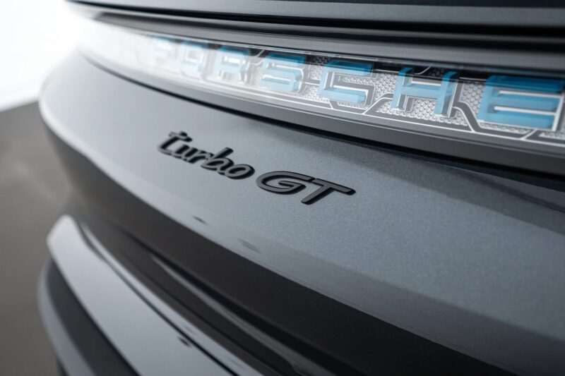 Close-up of the rear badge on a luxury car, highlighting the "turbo GT" badge and part of the illuminated "Porsche" emblem on a metallic gray surface.