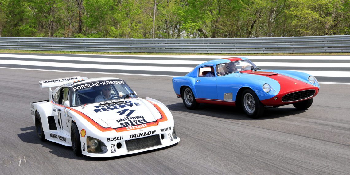A white Porsche race car and a blue classic Ferrari sports car drive side by side on a track with greenery in the background.