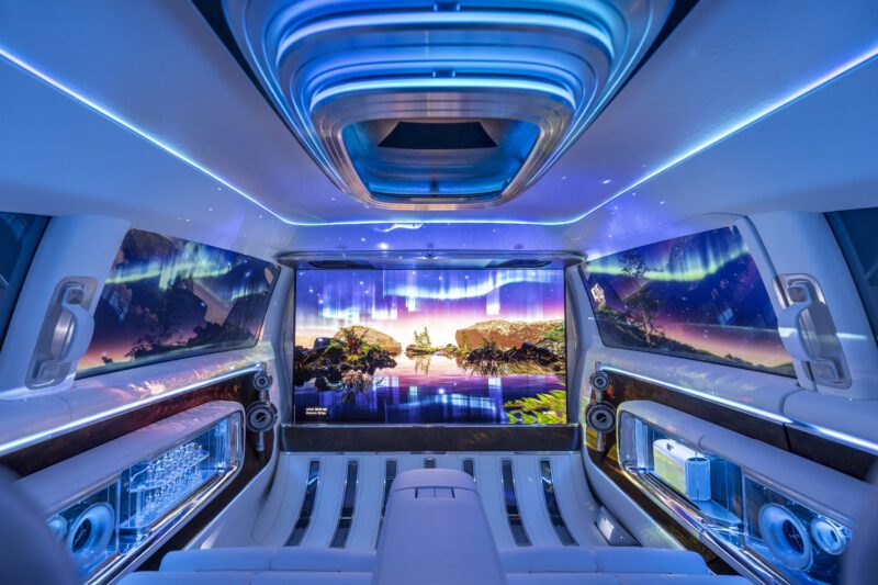 Luxury Mercedes-Benz Sprinter Van interior with blue ambient lighting, wide screen displaying northern lights, and elegant glassware storage on the side panels—welcome to the next level of comfort in the Vision V.