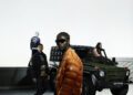 Four individuals in stylish clothing from the Moncler Collection stand on a rooftop beside a green off-road vehicle with "Past Future" on its grille. The backdrop is a clear overcast sky, perfectly complementing their fashion-forward presence.