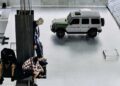 Three people sit near industrial structures on a metal platform, with a green and silver Mercedes-Benz SUV parked in the background.