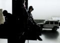 People in dark clothing sit in the foreground, with one wearing a NIGO-designed bucket hat. A sleek Mercedes-Benz SUV is parked in the background against a minimalist setting.