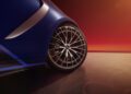 Close-up of a blue car's rear wheel with intricate rim design on a smooth surface, showcasing the luxury of the MANUFAKTUR Edition, against a gradient red-orange background.