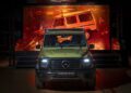 A green Mercedes-Benz G-Class SUV with headlights on is showcased indoors, standing before a brightly lit backdrop featuring an older G-Class model—highlighting how the new Mercedes-Benz is STRONGER THAN THE 1980s.