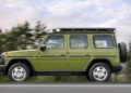 A 1-of-460 Mercedes-Benz G-Class, STRONGER THAN THE 1980s, with a roof rack and rear spare tire, drives on a road lined by trees beneath a cloudy sky.