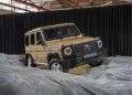 A tan Mercedes-Benz G-Class SUV, STRONGER THAN THE 1980s, is displayed on artificial rocky terrain indoors, set against black curtains and a ceiling of steel beams.