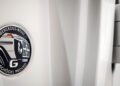 Close-up of a Mercedes-Benz G-Class "Schöckl Proved" badge on a white vehicle, hinting at its STRONGER THAN THE 1980s build, with a partial view of a door hinge on the right.