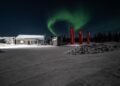 Under a starry night sky, the northern lights swirl over a snowy landscape. A cozy house with red banners stands in the foreground, resembling a Ferrari Purosangue in its elegance amid nature's spectacle.