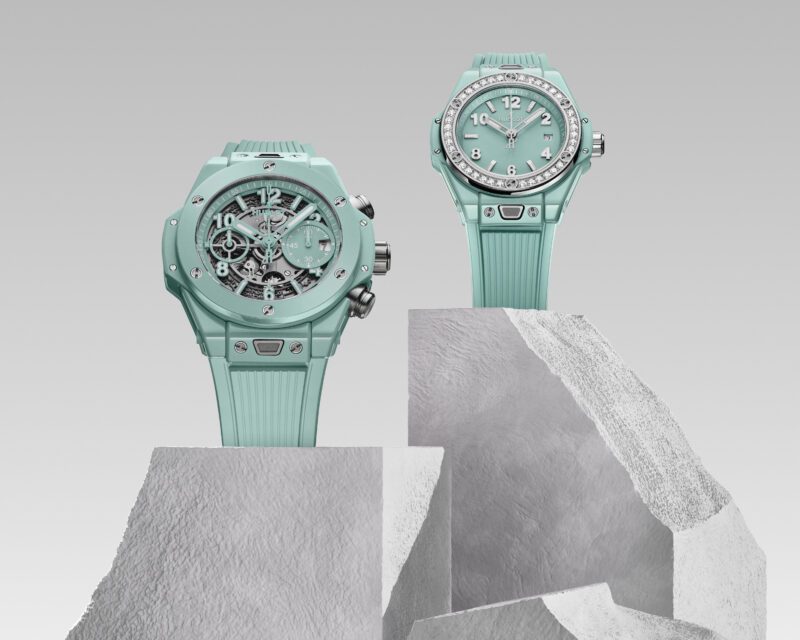 Two mint green Hublot watches with rubber straps gracefully rest on angular stone blocks. Celebrating the Big Bang's 20th Anniversary, one watch boasts an open skeleton design, while the other features a traditional face adorned with white numerals.