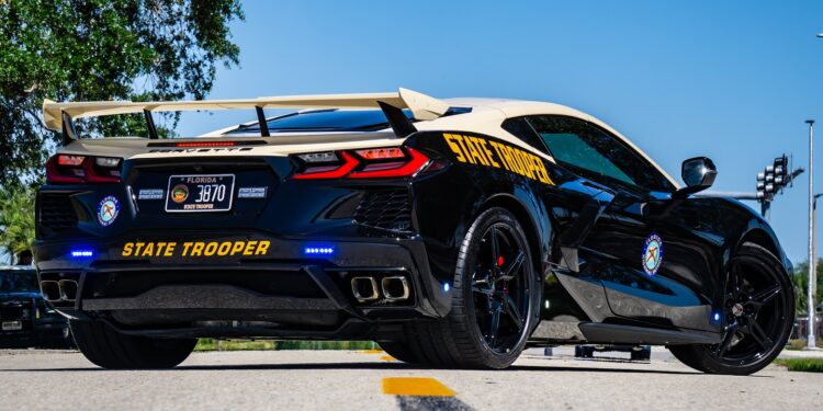 A sleek black and white C8 Corvette, adorned with a spoiler, emergency lights, and the Florida Highway Patrol insignia on its sides, is parked on a scenic road amidst trees under a clear blue sky.