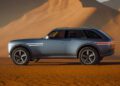 A blue Genesis X Gran Equator SUV with large wheels is parked on orange sand dunes under sunlight, casting shadows over the desert landscape in this striking off-road concept scene.