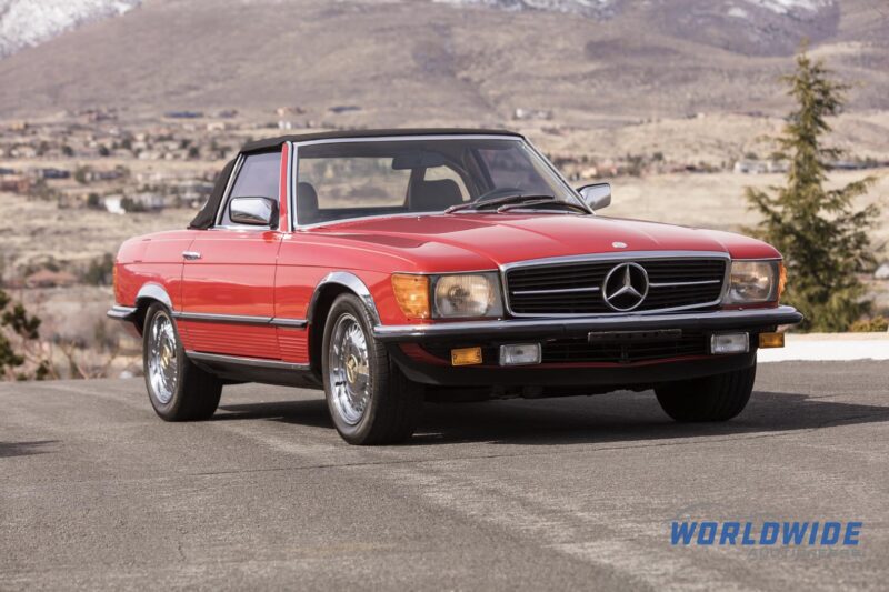 A red vintage Mercedes-Benz convertible is parked on a paved road with mountains and a town in the background, ready to turn heads at any Enthusiast Auction.
