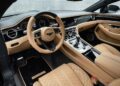 Luxury car interior with tan leather seats, wooden trim, and modern dashboard featuring a digital display, analog gauges, and various controls.