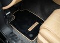 Black car floor mat with "MANSORY" branding placed in the driver's side footwell of a Bentley Continental GT featuring tan leather seats and carpeted flooring, highlighting refined customizations.