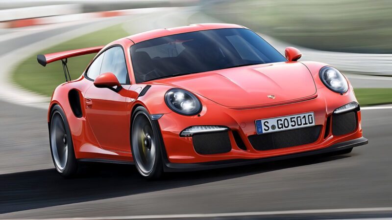 A red Porsche 911 GT3 RS with a rear wing is driving on a racetrack, captured from the front left side.